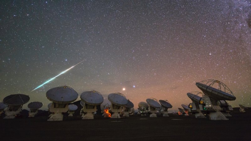 A bright light streaks across a starry, rosy-purple night sky on the left side of this image. An array of many radio telescopes are seen in the foreground.